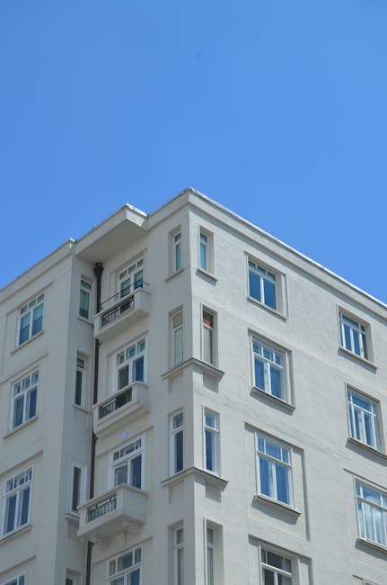 A row of white-fronted residential buildings with classic architectural details, featuring black wrought iron balconies decorated with hanging laundry and potted plants. In front, a paved street with