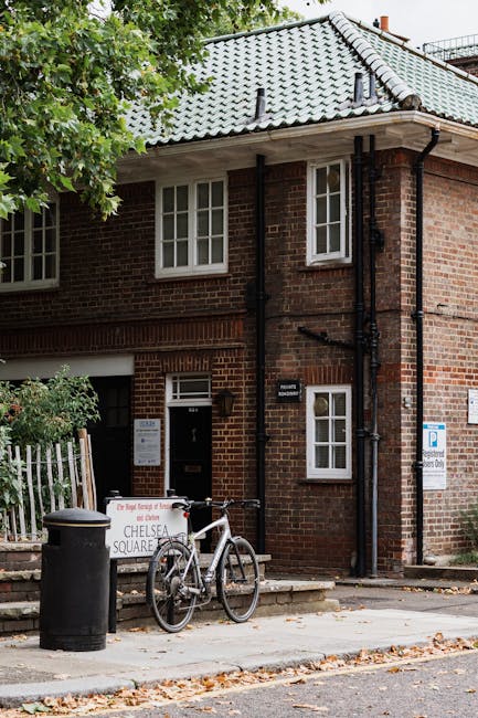 Photograph of a brick residential building with a grey tiled gable roof and white double-hung windows, situated on a street lined with trees. In front, a paved sidewalk features a black trash bin, a b